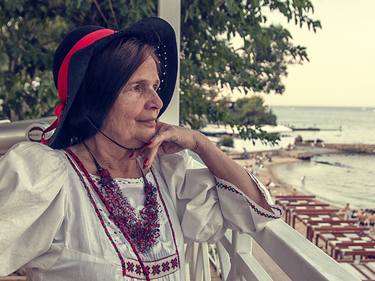 An old woman in a black hat, white dress and beads, looks from the balcony at the sea - Limited Edition of 3 thumb