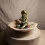 A sculpture made in away to express emottions through anarrative.an infand holding his milk in a shellwhere he feels safe though danger has already been in a short distance.glazed ceramic and acrylic paint used to show a childs figure,like a pearl in a shell damaged by the consequences of war